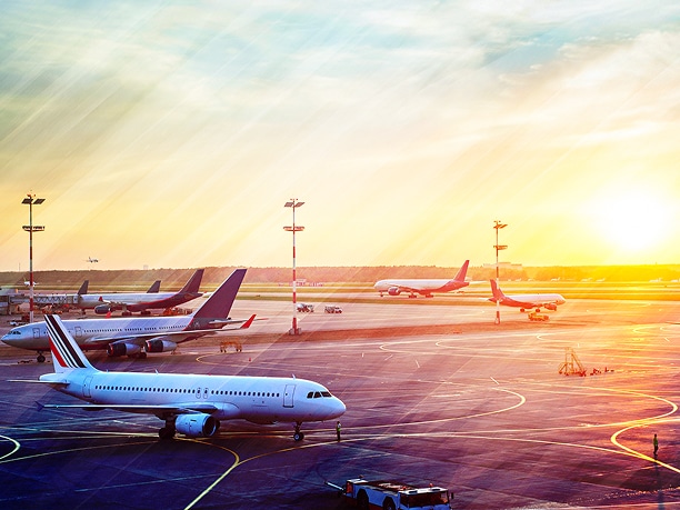Airplanes lined up at a busy airport terminal, preparing for takeoff at sunset.