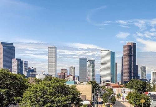 Vibrant skyline of Denver, Colorado with modern skyscrapers and trees in the foreground against a blue sky.