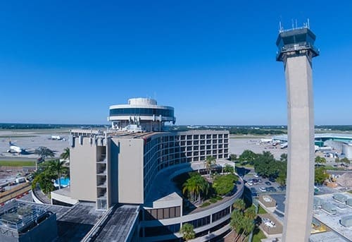 Aerial view of a large airport terminal and control tower against a clear blue sky.