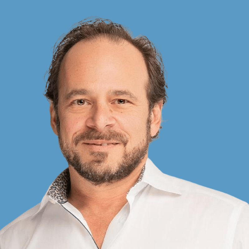 Portrait of a middle-aged man with brown hair and a beard, wearing a white button-up shirt and smiling against a blue background.