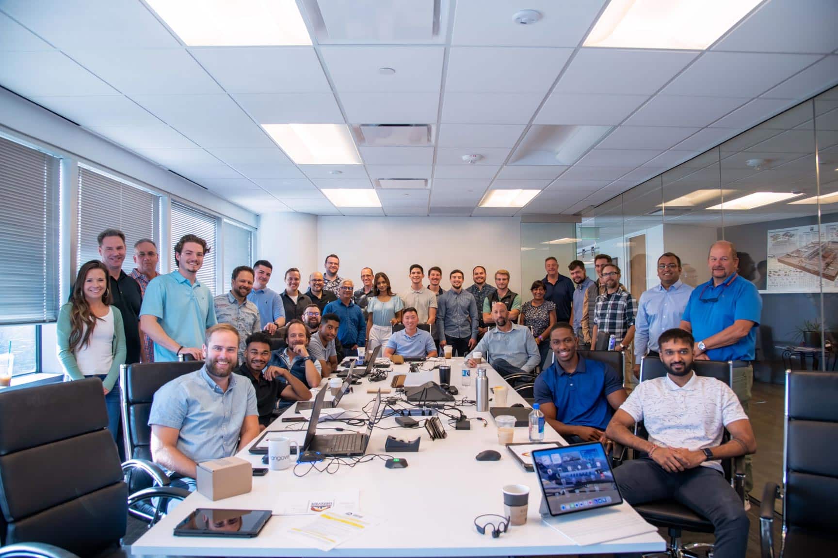 A large group of people, mostly men, gathered in an office setting with desks, computers, and other office equipment.