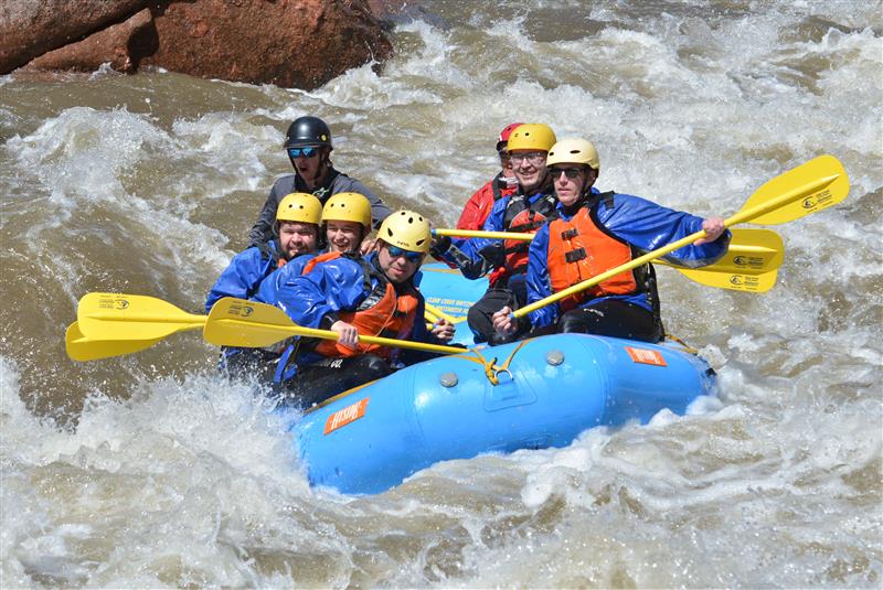 Group of people rafting through whitewater rapids, wearing safety gear.