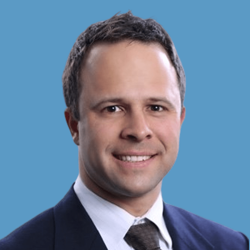 Smiling professional man with dark hair wearing a navy suit and tie against a blue background.