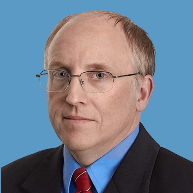 Middle-aged man in formal business attire with glasses, sitting in front of blue background.
