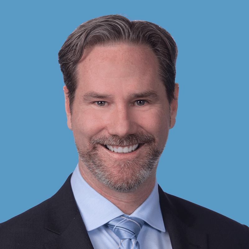 A smiling man with gray hair and beard wearing a suit and tie against a blue background.
