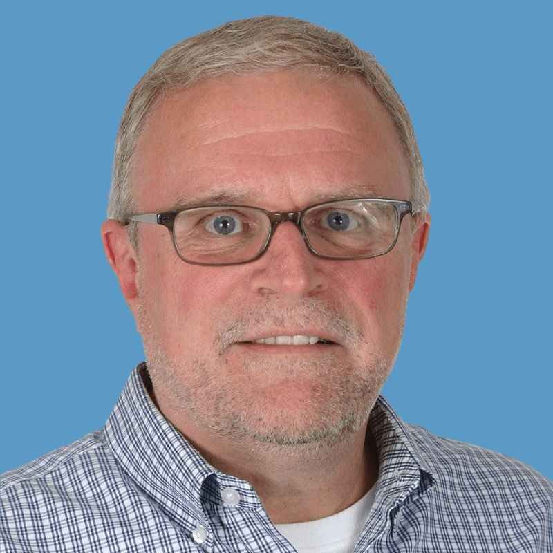 Smiling middle-aged man with gray hair and glasses wearing a plaid shirt against a blue background.