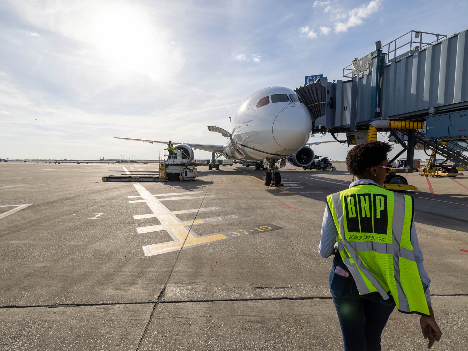 Airplane on tarmac beside airport jetway and ground crew member in high-visibility vest.